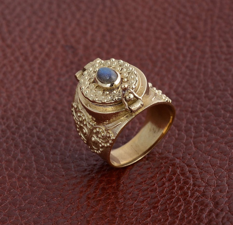 Puede incluir: Un anillo de oro con un compartimento con bisagras y una piedra preciosa azul engastada en la parte superior. El anillo est&aacute; intrincadamente detallado con patrones florales.
