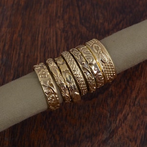 May include: A stack of eight gold rings with intricate floral and geometric designs. The rings are stacked on a brown surface.