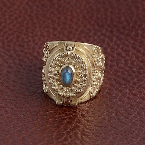 Puede incluir: Un anillo de oro con una piedra preciosa azul engastada en un dise&ntilde;o circular. El anillo tiene una abertura con bisagra y detalles intrincados.