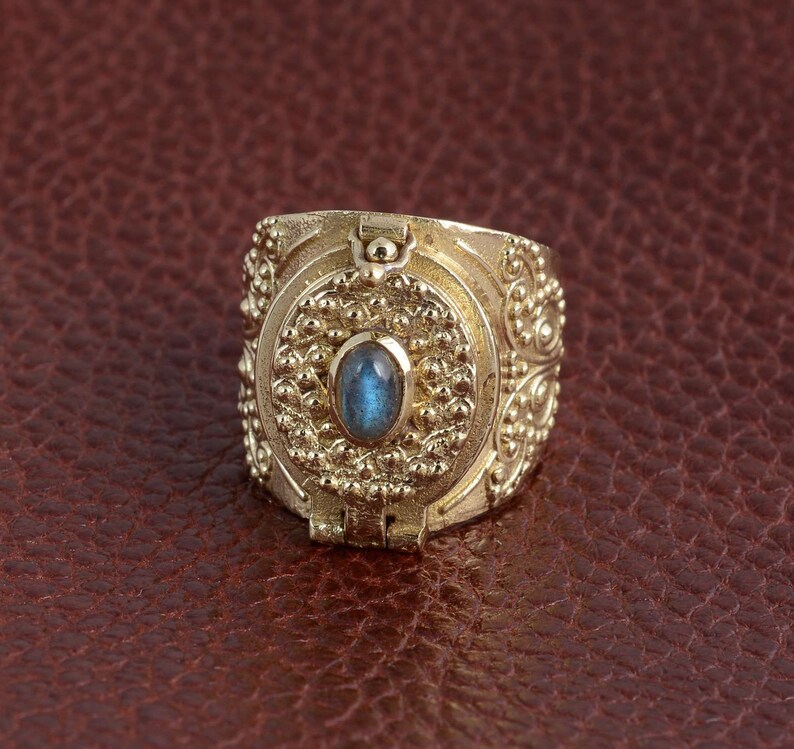 Puede incluir: Un anillo de oro con una piedra preciosa azul engastada en un dise&ntilde;o circular. El anillo tiene una abertura con bisagra y detalles intrincados.