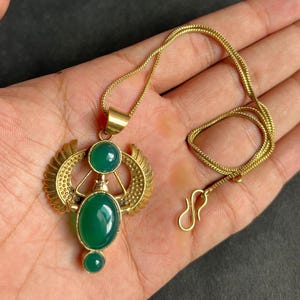 以下が含まれることがあります： 緑色の石のペンダントがついた金のネックレス。翼のある女神のデザインです。