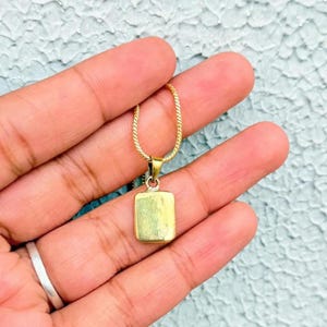 Könnte beinhalten: Eine goldfarbene Halskette mit einem rechteckigen Anhänger. Der Anhänger hängt an einer filigranen Kette. Die Halskette wird in einer Hand vor einem strukturierten, hellblauen Hintergrund gehalten.