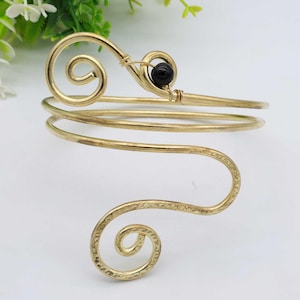 Könnte beinhalten: Ein goldfarbenes Metallarmband mit einem schwarzen Perlenakzent. Das Armband besteht aus Draht, der zu einem Spiraldesign verdreht ist.