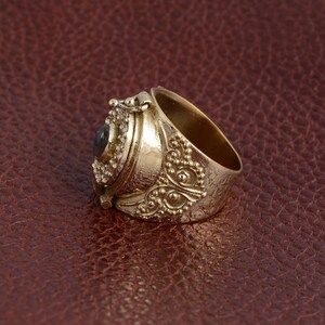 Puede incluir: Un anillo de oro con un compartimento con bisagras y una peque&ntilde;a piedra azul oscuro. El anillo est&aacute; decorado con intrincados patrones florales y peque&ntilde;as cuentas.