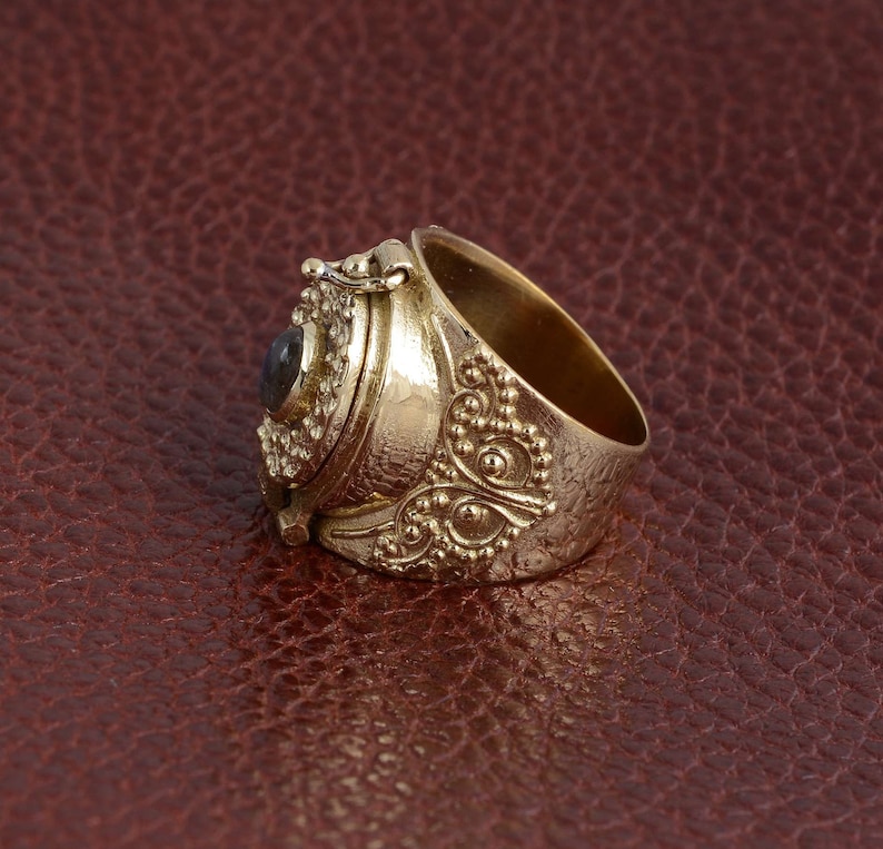 Puede incluir: Un anillo de oro con un compartimento con bisagras y una peque&ntilde;a piedra azul oscuro. El anillo est&aacute; decorado con intrincados patrones florales y peque&ntilde;as cuentas.