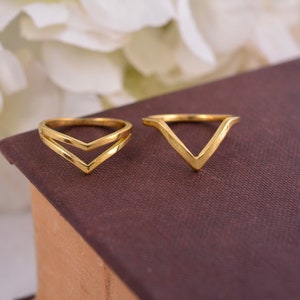 Puede incluir: Dos anillos dorados con un diseño en forma de V. El anillo de la izquierda tiene una doble forma de V, mientras que el anillo de la derecha tiene una sola forma de V.