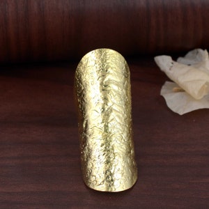 May include: A gold-toned, textured metal ring with a wide band. The ring is designed to be worn on the knuckle.