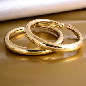 Könnte beinhalten: Zwei goldfarbene Armreifen, einer etwas größer als der andere, liegen übereinander. Die Armreifen sind glatt und glänzend.