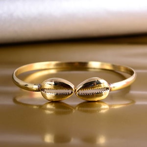 May include: A gold-toned bangle bracelet featuring two shell-shaped charms. The bracelet is simple and elegant, ideal for everyday wear.