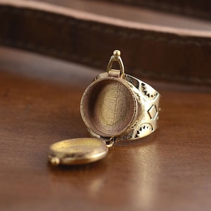 May include: A gold-toned ring featuring a hinged, round locket that opens to reveal an empty interior. The ring band is decorated with a geometric pattern.