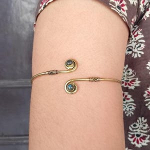 以下が含まれることがあります： 2つの青緑色の宝石のアクセントが付いた金の腕輪。腕輪はワイヤー製で、シンプルでエレガントなデザインです。