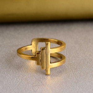 Puede incluir: Un anillo de color dorado con un diseño geométrico. El anillo está hecho de dos bandas que están conectadas por una forma cuadrada con una forma cuadrada más pequeña en la parte superior.