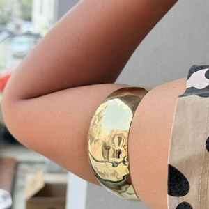 May include: A gold-toned metal cuff bracelet with a smooth, rounded surface. The bracelet is worn on the upper arm.