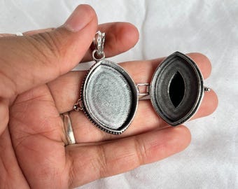 Op de afbeelding: Open zilverkleurige locket hanger met een ovale vorm. De locket is open en onthult aan de ene kant een zwarte steen en aan de andere kant een getextureerde, lege ruimte. De hanger wordt in een hand gehouden.
