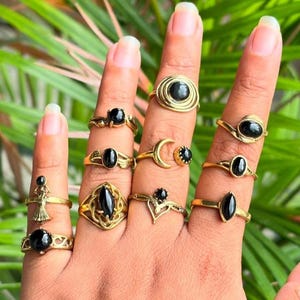 May include: A collection of ten gold-toned rings featuring black oval, round, and marquise-cut stones. The designs include a crescent moon, a figure-shaped setting, intricate wirework, and simple bands, all worn across four fingers.