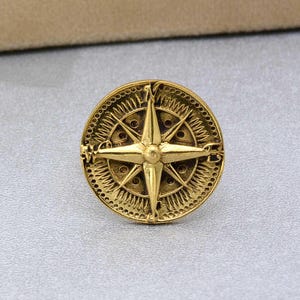 May include: A gold-toned compass with a detailed design featuring a star-shaped compass rose and a circular border.