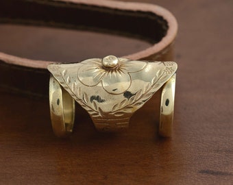 Pode incluir: Um anel de metal dourado com uma placa superior larga e curva gravada com um motivo floral central e padrões de folhas. A banda consiste em dois aros circulares separados conectados pela placa decorativa, criando uma estrutura singular.