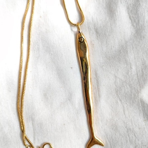 Op de afbeelding: Een gouden ketting met een gouden vis-hanger. De vis is lang en slank met een klein oog.