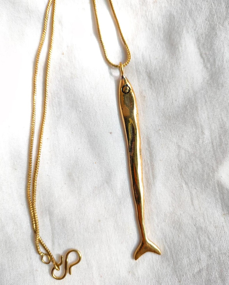 Op de afbeelding: Een gouden ketting met een gouden vis-hanger. De vis is lang en slank met een klein oog.