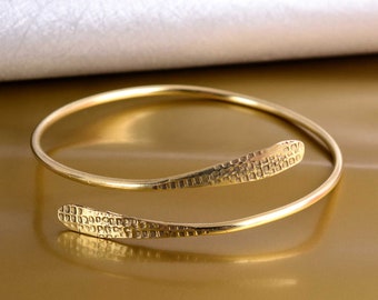 May include: A gold-toned bangle bracelet with a textured, patterned design. The bracelet is open and has a simple, minimalist style.