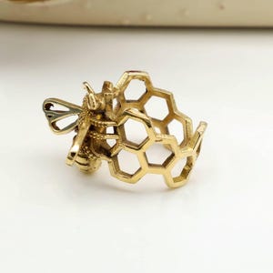 Op de afbeelding: Een goudkleurige ring met een gedetailleerd ontwerp van een bij en een honingraat. De ring heeft een gedetailleerde bijenfiguur en een honingraatvormige band. De vleugels en het lichaam van de bij zijn ingewikkeld ontworpen.