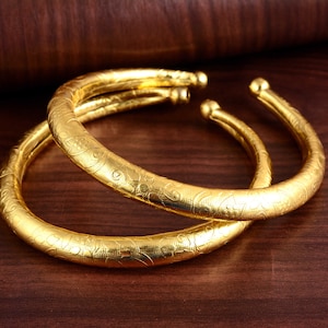 May include: Two gold bangles with intricate floral designs. The bangles are stacked on top of each other.