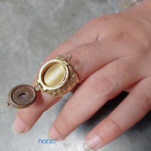 Puede incluir: Un anillo de oro con un compartimento redondo adornado con bisagras que se abre para revelar una peque&ntilde;a piedra redonda de color p&uacute;rpura. El anillo se lleva en un dedo.