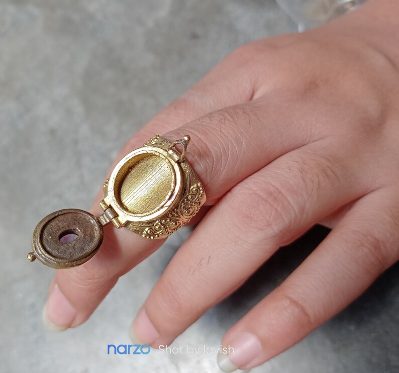 Puede incluir: Un anillo de oro con un compartimento redondo adornado con bisagras que se abre para revelar una peque&ntilde;a piedra redonda de color p&uacute;rpura. El anillo se lleva en un dedo.