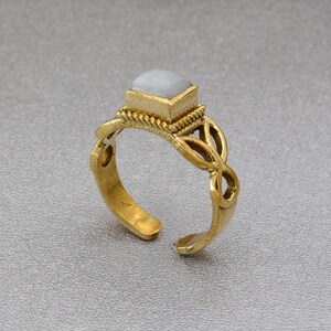 May include: A gold ring with a white stone set in a square shape. The ring has a delicate, intricate design with a Celtic knot pattern.