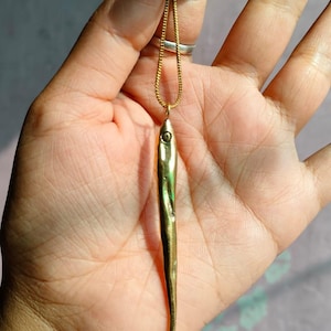 Op de afbeelding: Een gouden ketting met een lange, dunne, gouden vis-hanger.