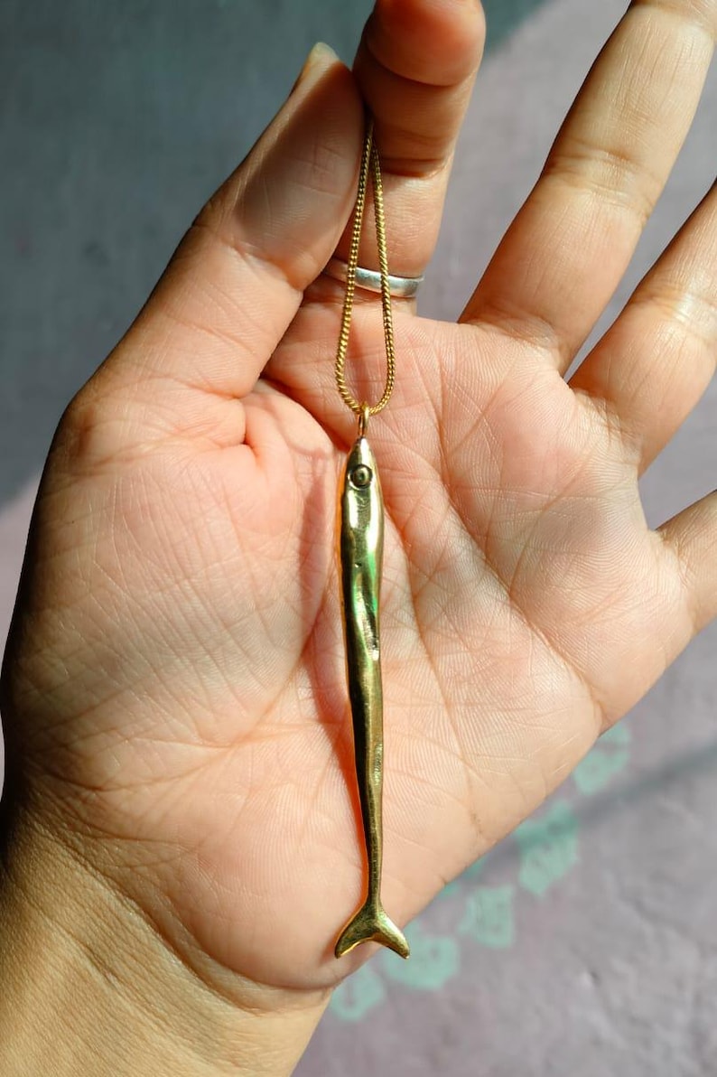 Op de afbeelding: Een gouden ketting met een lange, dunne, gouden vis-hanger.