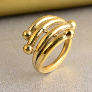 May include: A gold ring with a unique design featuring three intertwined bands with rounded ends. The ring is made of a smooth, polished metal.