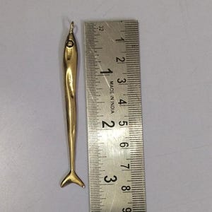Op de afbeelding: Een gouden vis-hanger, ongeveer 5 cm lang, met een lus aan de bovenkant om aan een ketting of armband te bevestigen. De hanger is gedetailleerd met een getextureerd oppervlak en een gevorkte staart.