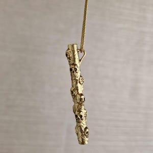 May include: A gold chain necklace with a pendant shaped like a long, thin branch with multiple skull-shaped carvings.