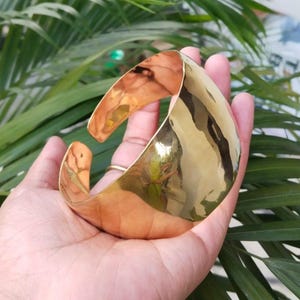 May include: A wide, open-cuff bracelet with a polished, golden finish. The cuff is made of a thick metal and has a smooth, reflective surface. The bracelet is held in a hand, with green foliage in the background.