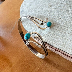 Puede incluir: Brazalete plateado con dos piedras de color turquesa. El brazalete presenta un diseño abierto con remolinos decorativos y un acabado pulido. Las piedras turquesas son de forma ovalada y están engastadas en un bisel. El brazalete se exhibe sobre una superficie de madera.