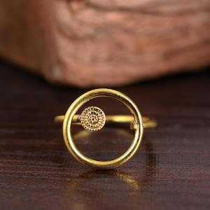Könnte beinhalten: Ein goldener Ring mit einem Kreisdesign. Der Kreis ist offen und hat einen kleinen, strukturierten Kreis in der Mitte.