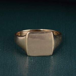 Puede incluir: Un anillo de sello de oro con una parte superior rectangular en blanco. El anillo está sobre una superficie verde oscuro.