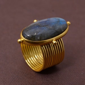 Puede incluir: Un anillo de oro con una gran piedra ovalada de labradorita azul engastada en un engaste de bisel. La banda del anillo está hecha de múltiples bandas finas de oro.
