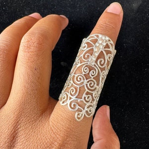 May include: A silver filigree ring with a delicate swirl design. The ring is wide and sits high on the finger.