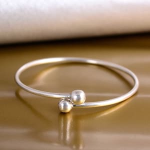May include: A silver-colored bangle bracelet with two small, round, decorative balls. The bracelet has a simple, elegant design and is set against a gold-toned background. The bracelet is a piece of jewelry.