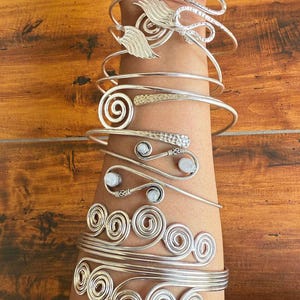 May include: A set of silver bracelets with a variety of designs, including a spiral, a wing, and a snake. The bracelets are stacked on a person's arm.
