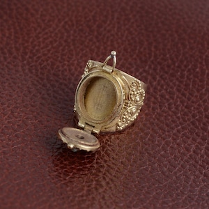 Puede incluir: Un anillo dorado con una tapa con bisagras que se abre para revelar un peque&ntilde;o compartimento redondo. El anillo est&aacute; decorado con intrincados patrones florales y peque&ntilde;as cuentas.