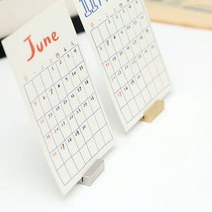 May include: Two white calendar cards with blue grid lines and black numbers. The calendar cards are standing upright on small metal and wooden stands. The calendar cards are for June and July.