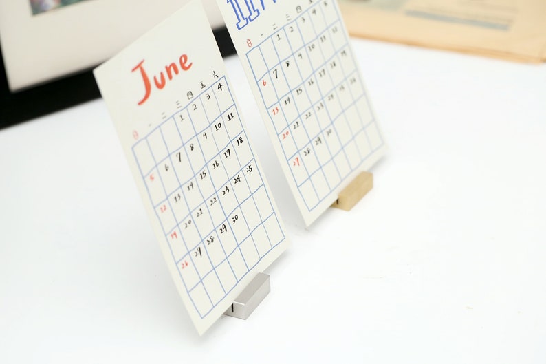 May include: Two white calendar cards with blue grid lines and black numbers. The calendar cards are standing upright on small metal and wooden stands. The calendar cards are for June and July.