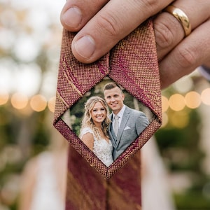 Puede incluir: Una corbata burdeos y dorada con un estampado y una foto de una pareja sonriente. La corbata está sujeta por una mano, con un anillo de bodas dorado visible. La foto está enmarcada por la tela de la corbata.