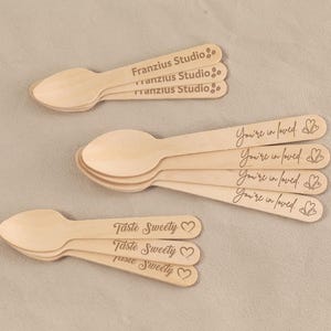May include: Wooden spoons with engraved text. Some spoons read "Franzius Studio," others say "You're in loved" with a heart, and some say "Taste Sweety" with a heart. The spoons are light brown.