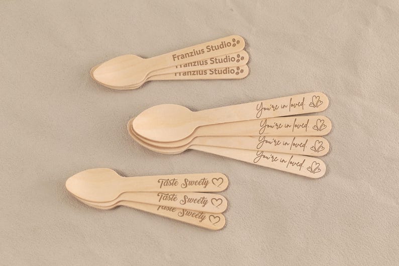 May include: Wooden spoons with engraved text. Some spoons read "Franzius Studio," others say "You're in loved" with a heart, and some say "Taste Sweety" with a heart. The spoons are light brown.