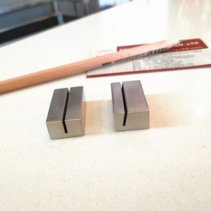 May include: Two silver metal business card holders with a slot for cards. The holders are on a white surface.