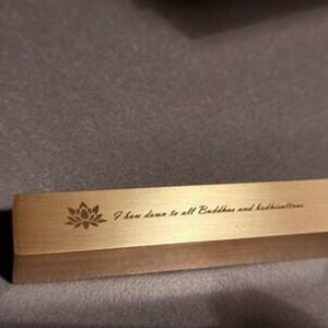 May include: A gold-colored metal bar with a lotus flower design and the text "I bow down to all Buddhas and bodhisattvas".